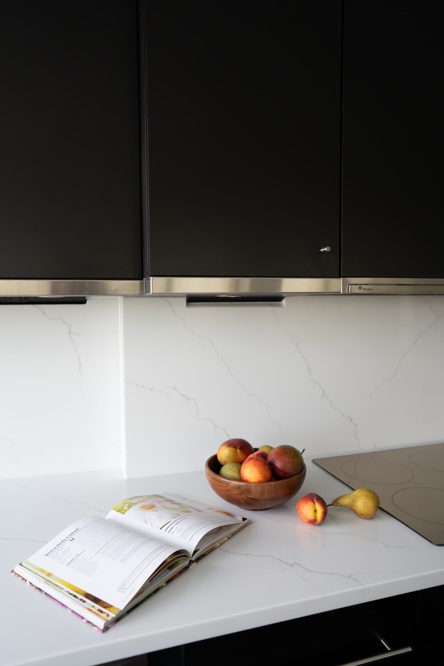 Kitchen vignette: a wood bowl of fruit and an open cookbook on the white quartz counter beneath matte-black cabinets.