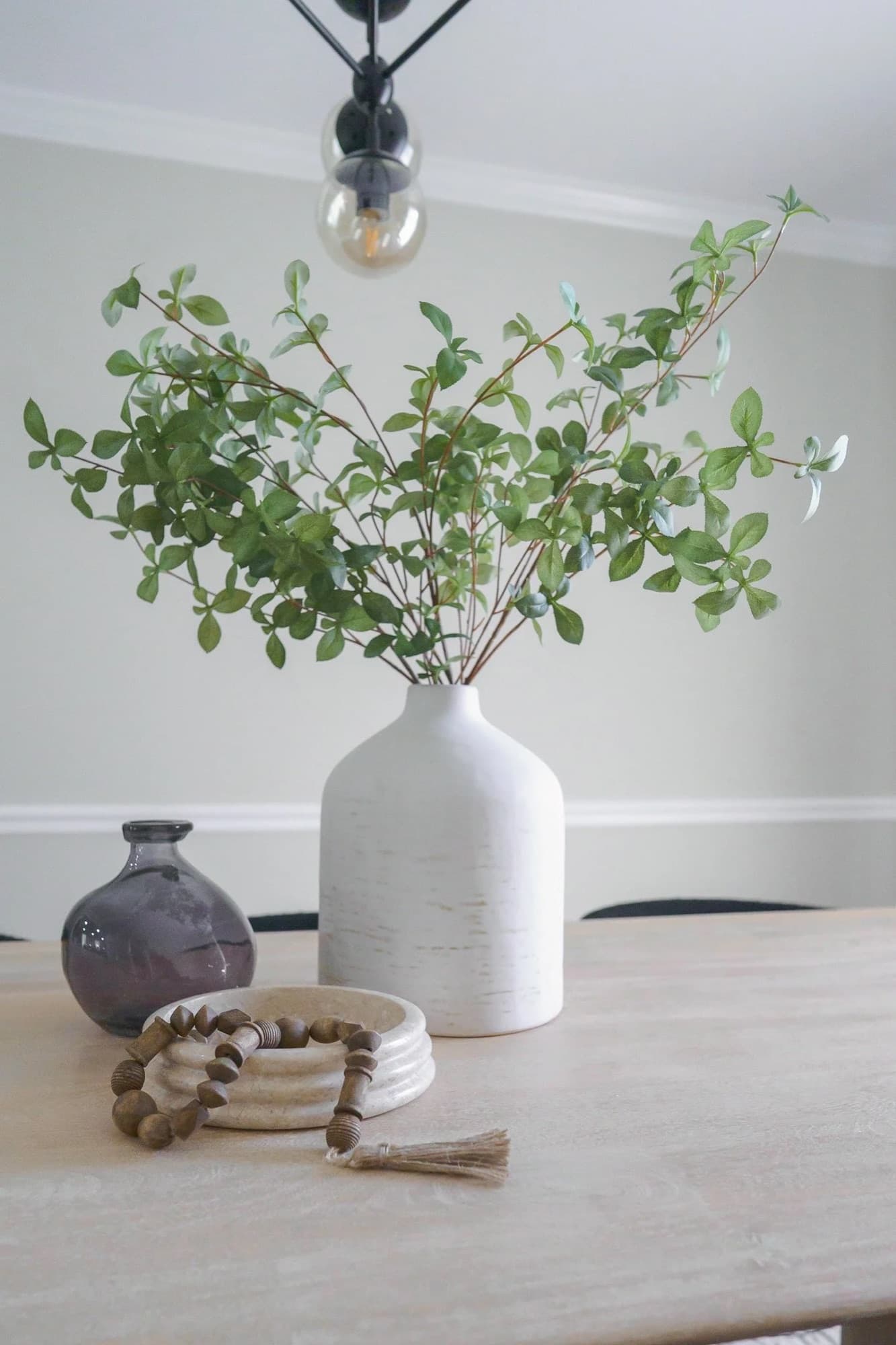 Dining table centerpiece: a white textured vase of greenery beside a smoky glass bud vase and a wood bead detail.