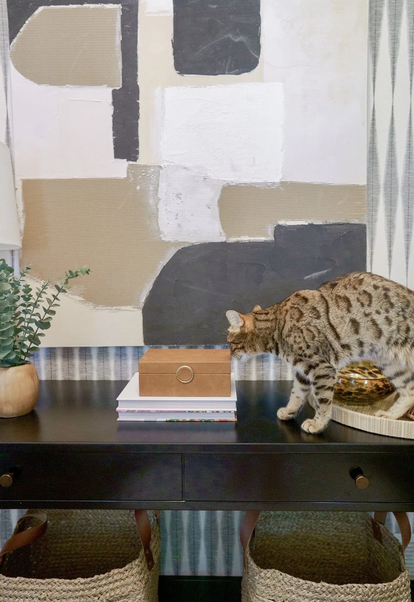 Entry vignette with a black console table, layered abstract art, woven baskets, and the family's cat mid-step.
