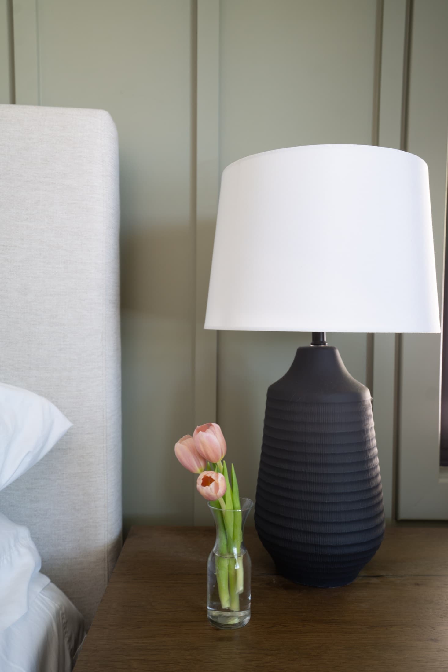 Bedside detail: a ribbed navy lamp and a single pink tulip in a bud vase on a wood nightstand.