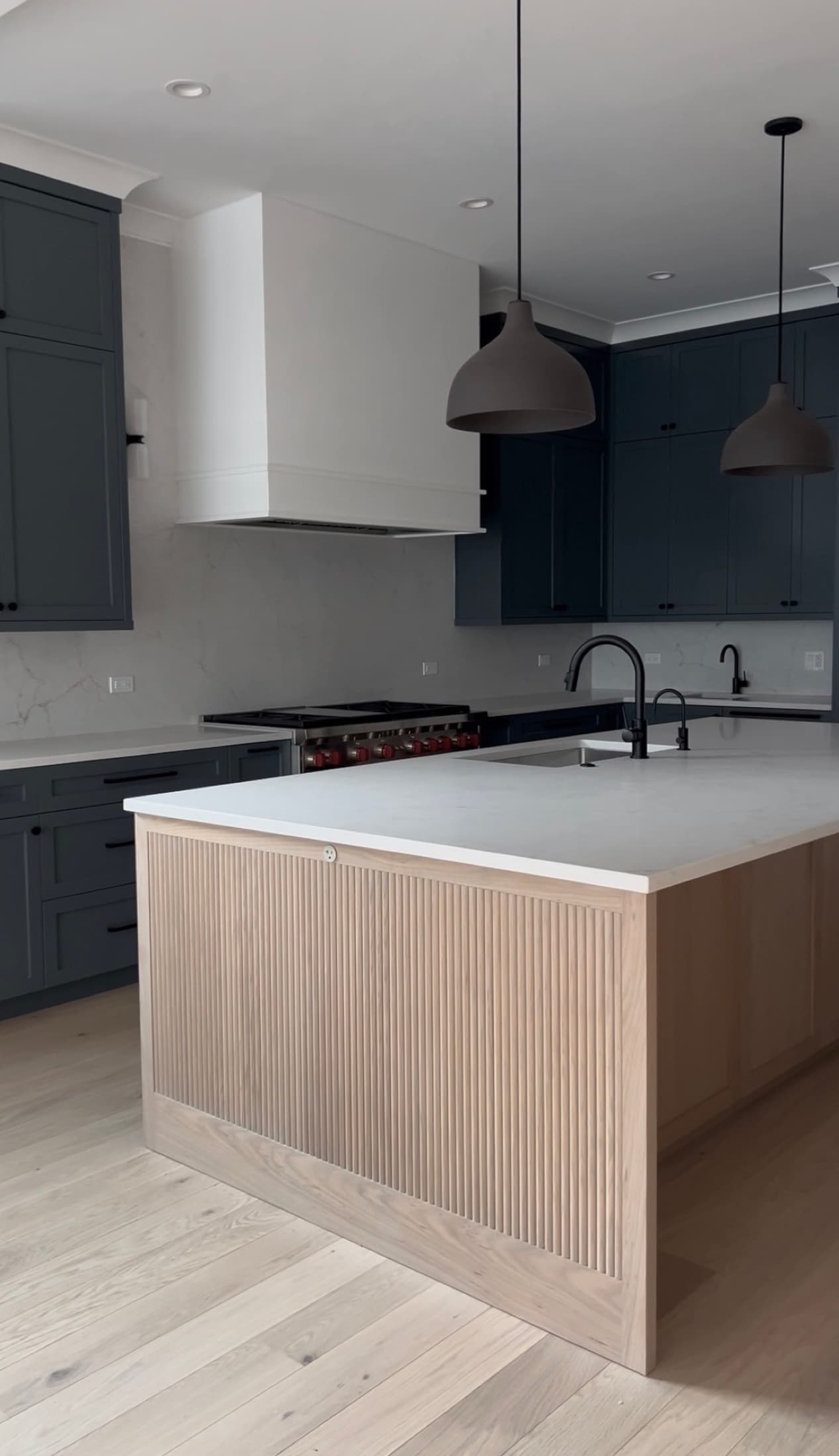 Superior Living kitchen with slate-blue cabinetry, a white-oak fluted island, marble backsplash, and black dome pendants over herringbone wood floors.