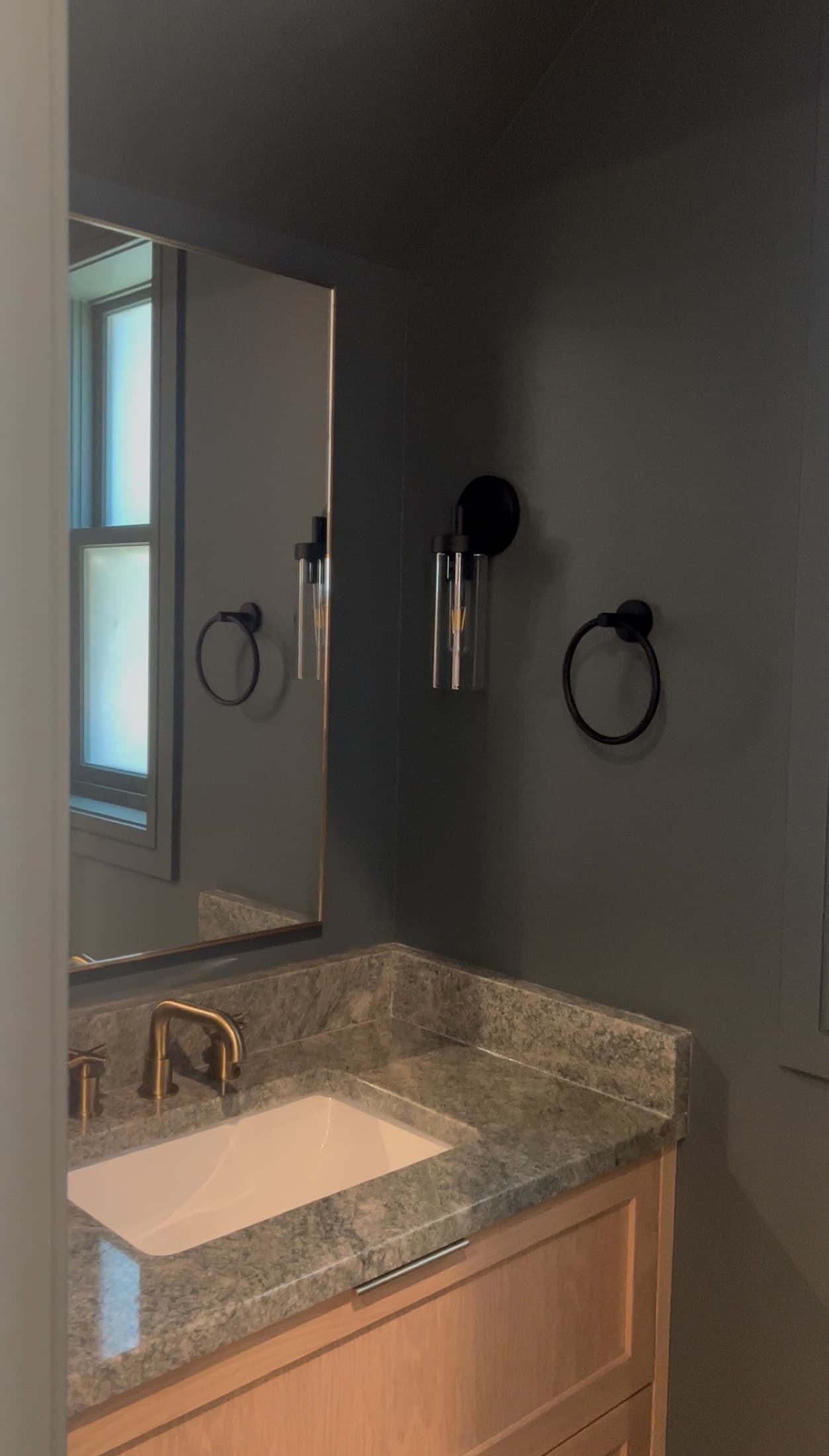 Powder bath with warm gray walls, a marble-topped light-wood vanity, brushed-brass faucet, and a seeded-glass sconce.